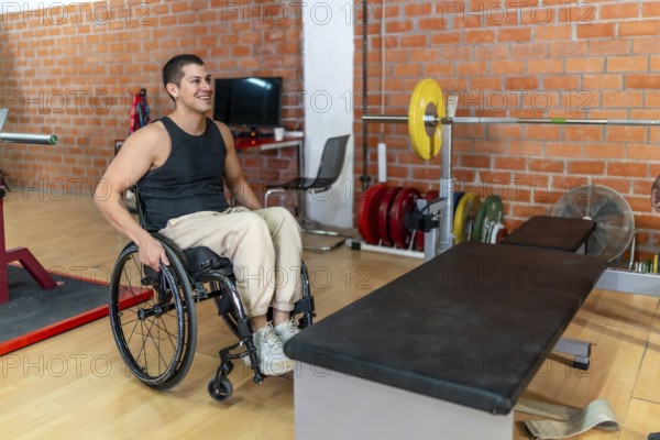Young man in a wheelchair preparing for a workout next to a weightlifting bench and barbell at an indoor gym, focusing on adaptive fitness and inclusion