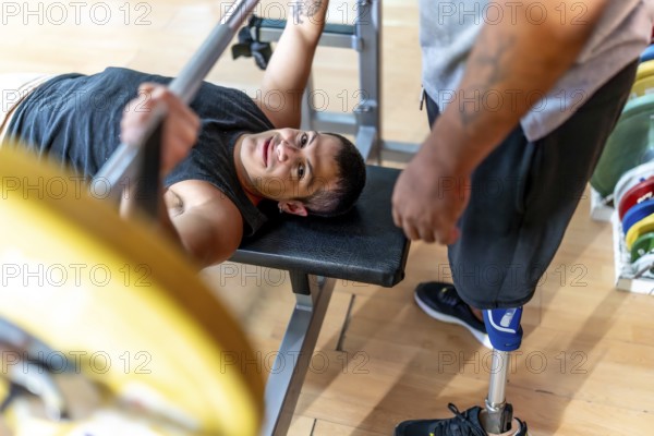 Disabled man performing a bench press with a barbell while a friend with a prosthetic leg stands by, offering support during a workout session at the gym