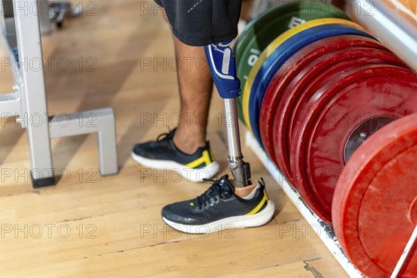 Amputee man standing on a wooden floor in a gym, wearing a modern prosthetic leg and a normal shoe on the other foot, next to a rack of colorful weight plates