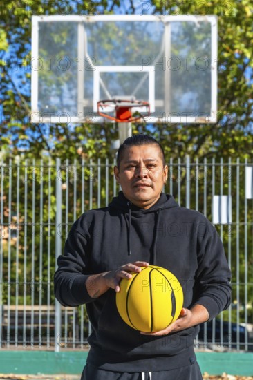 Indigenous man standing on an outdoor basketball court holding a bright yellow basketball, looking seriously at the viewer on a sunny day with a hoop in the background