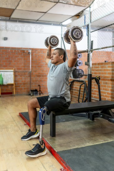 Man with a prosthetic leg performing overhead dumbbell presses while sitting on a weight bench, focusing on upper body strength and adaptive fitness in a gym environment