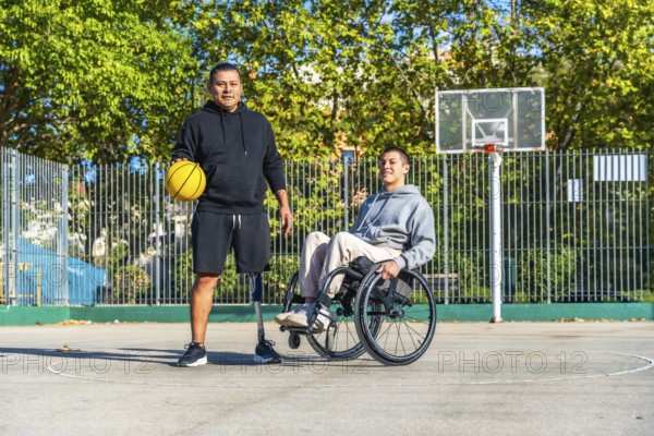Two young men, one with a prosthetic leg holding a basketball and another sitting in a wheelchair, are standing together on an outdoor basketball court, representing inclusion and adaptive sports