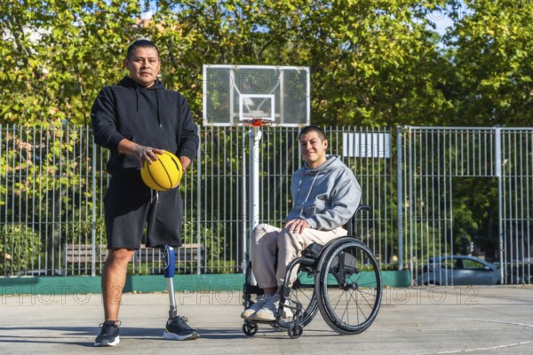 Two young men, one with a prosthetic leg holding a basketball and another sitting in a wheelchair, standing together on an outdoor basketball court in a park, representing adaptive sports