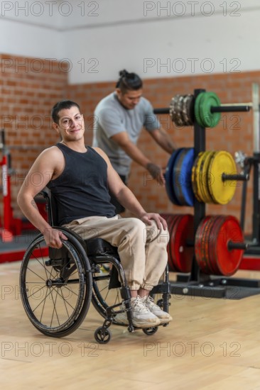Disabled man in a wheelchair looking cheerful and smiling at the camera while another man with a prosthetic leg is preparing weights in a gym, showing inclusivity and adaptive fitness