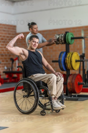 Adaptive athlete in a wheelchair flexing his bicep and smiling while a trainer adjusts weight equipment behind him in a bright gym, showing strength and determination