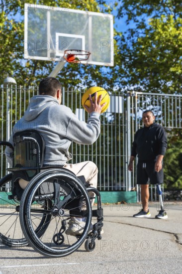 Man in a wheelchair aiming to shoot a basketball while his male friend, who uses a prosthetic leg, stands watching on an outdoor basketball court, representing inclusion and adapted sports