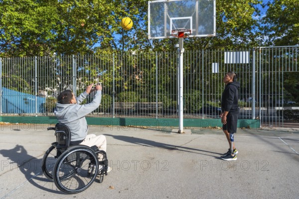 Two friends with disabilities play basketball outdoors, one shoots from a wheelchair while the other with a prosthetic leg defends, sharing adaptive sport, teamwork, joy and determination
