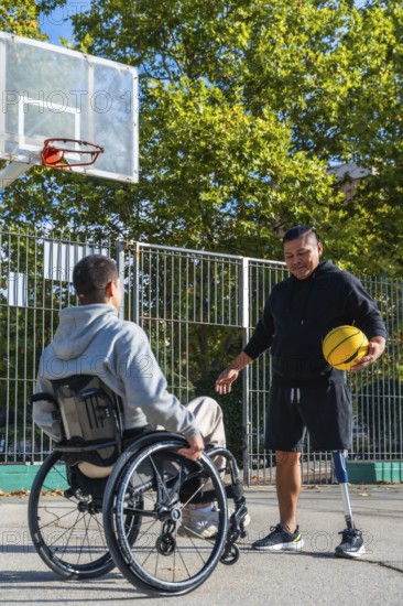 Two disabled men are enjoying a game of basketball on an outdoor court, with one man using a wheelchair and the other having a prosthetic leg, symbolizing inclusion and sport