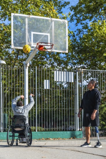 Two men play outdoor basketball, one shooting from a sports wheelchair while the other stands with a prosthetic leg, showcasing adaptive sport, teamwork and friendship on court