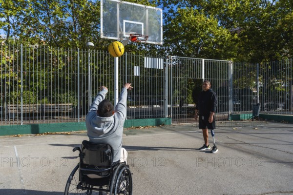 Two young men playing basketball outdoors, one in a wheelchair shooting the ball towards the hoop and the other standing with his prosthetic leg, showing friendship, inclusion, and adaptive sports