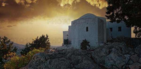 The white chapel of the Virgin Mary Tsambika (Kyra Psili) in front of a dramatic sunset sky with rocks and trees, Church of the Virgin Mary Tsambika (Kyra Psili), Archangelos, Rhodes Island, Greece