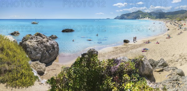 Picturesque Tsambika bay with clear water, rocks and sandy beach. People enjoying the sun, a boat in the background, lush vegetation, Tsambika beach, Archaggelos, Rhodes island, Greece
