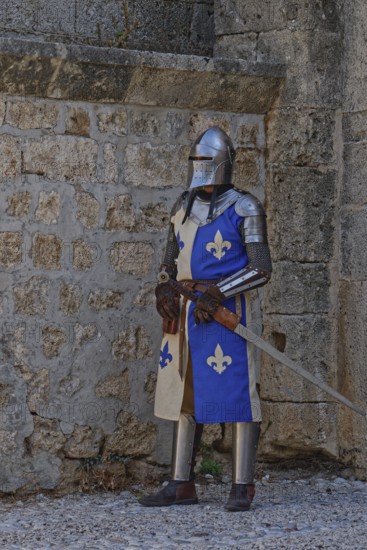A knight in medieval armour stands with a sword in front of a stone wall, Medieval Town of Rhodes, Rhodes Island, Greece