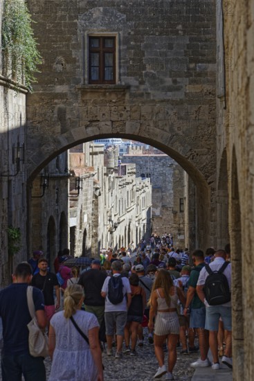 A busy historic alleyway with people walking through an alley under a stone arch, Knights Street, Medieval Town of Rhodes, Rhodes Island, Greece