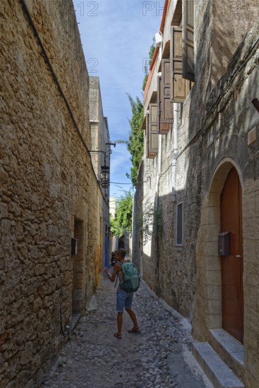 A woman with a rucksack walks through a narrow cobbled alley between historic stone houses, Medieval Town of Rhodes, Rhodes Island, Greece