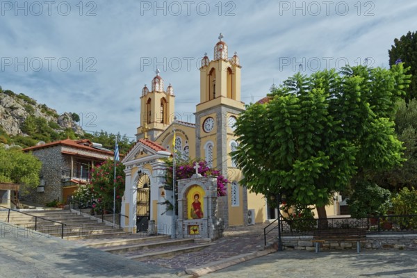 A yellow church with towers and bells surrounded by trees under a cloudy sky, Saint Pantaleon Greek Orthodox Church, Siana, Rhodes Island, Greece