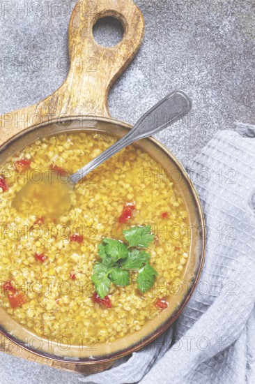 A nourishing bowl of lentil soup filled with grains and vibrant red pepper pieces sits on a textured gray countertop. Fresh herbs add a touch of color and flavor