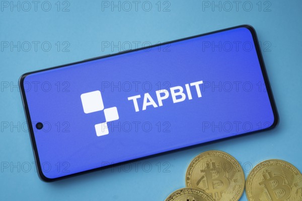 Dhaka, Bangladesh- 19 Sep 2025: Tapbit logo on smartphone screen near Bitcoin coin and QR code on blue background