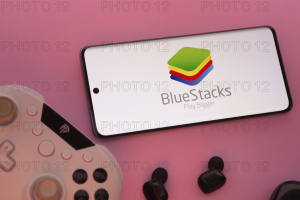Dhaka, Bangladesh- 23 Sep 2025: Smartphone with BlueStacks logo, game controller, and earbuds on a pink background
