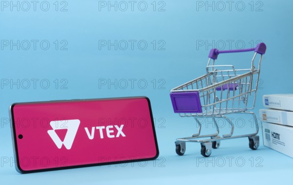Dhaka, Bangladesh- 03 July 2025: Smartphone with VTEX logo next to a small shopping cart and boxes against a blue background
