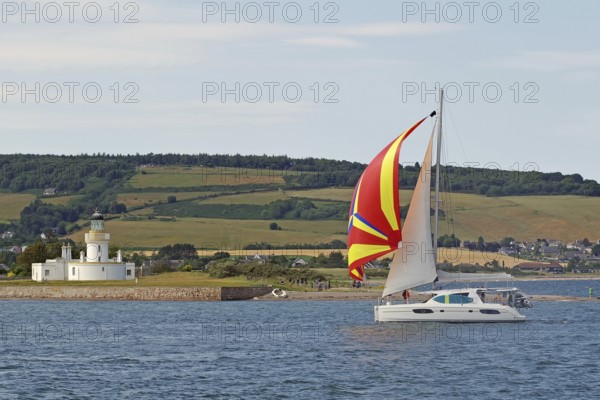 A sailing boat with a colourful sail sails near a lighthouse on the coast, Moray Firth, Inverness, Scotland, Great Britain