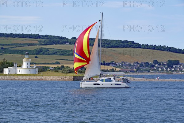 A sailing boat with a coloured sail sails near a lighthouse in front of a green landscape, Moray Firth, Inverness, Scotland, Great Britain