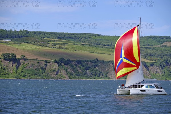 A sailing boat with a vividly coloured flag sails in front of a mountainous coastal landscape, Moray Firth, Inverness, Scotland, Great Britain