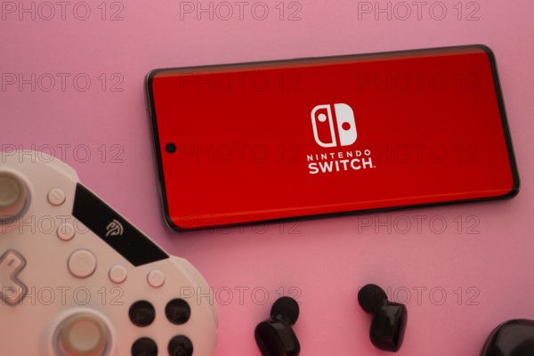Dhaka, Bangladesh- 15 July 2025: Smartphone with nintendo switch logo, game controller, and earbuds on a pink background