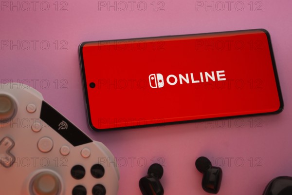 Dhaka, Bangladesh- 15 July 2025: Smartphone with nintendo online logo, game controller, and earbuds on a pink background