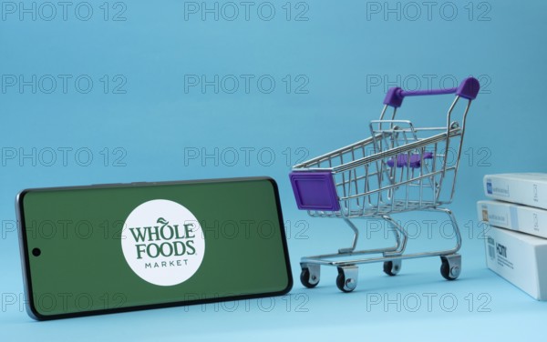 Dhaka, Bangladesh- 03 July 2025: Smartphone with Whole Foods Market logo next to a small shopping cart and boxes against a blue background