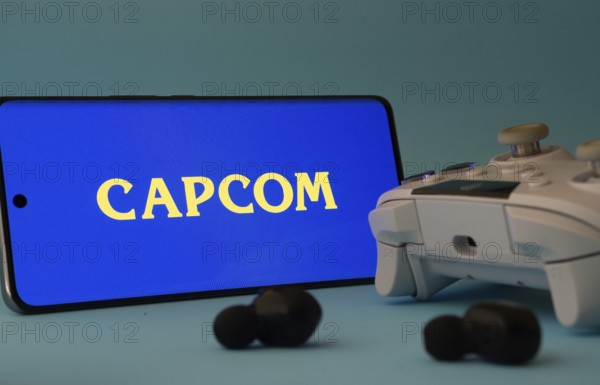 Dhaka, Bangladesh- 19 Sep 2025: A smartphone with a Capcom icon next to a controller and earbuds on a blue surface