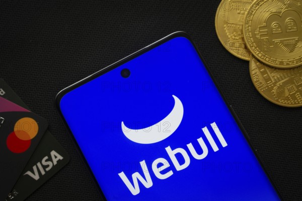 Dhaka, Bangladesh- 26 Aug 2025: Smartphone displaying webull logo surrounded by Bitcoin coins and payment cards