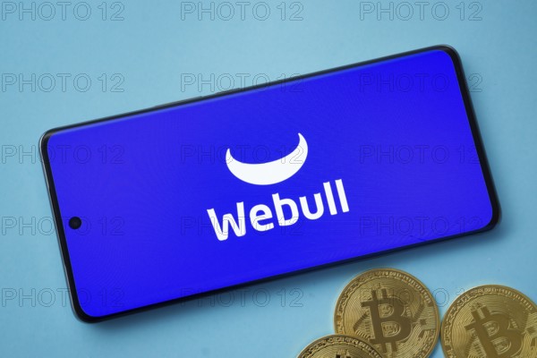 Dhaka, Bangladesh- 03 Sep 2025: webull logo on smartphone screen near Bitcoin coin and QR code on blue background