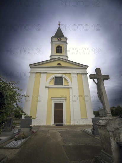Yellow church with tower and cross in a cemetery under a cloudy sky, Kammersdorf Hollabrunn Weinviertel Lower Austria Austria