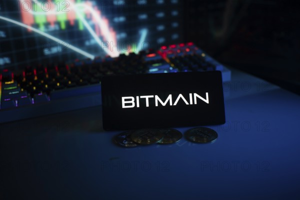 Dark-themed Bitmain logo on a phone with coins and trading screen background
