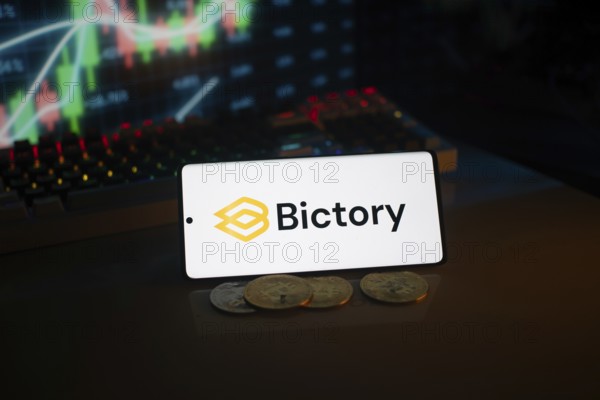 Bictory logo on smartphone amidst coins and stock chart, conveying digital and financial themes