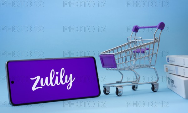 Dhaka, Bangladesh- 03 July 2025: Smartphone with Zulily logo next to a small shopping cart and boxes against a blue background