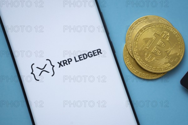 Dhaka, Bangladesh- 26 Aug 2025: XRP Ledger logo on smartphone screen near Bitcoin coin and QR code on blue background