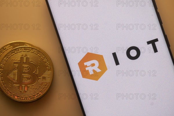 Bitcoin coin near phone displaying RIOT logo on a white background