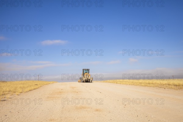 In the remote desert near Solitaire, Namibia, a tractor levels a dirt road across golden sand dunes and rugged desert terrain