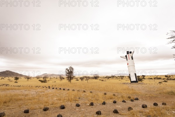 A mock Soviet rocket stands nose-first in the Solitaire, Namibian desert, surrounded by golden grasslands and rugged mountains under cloudy skies