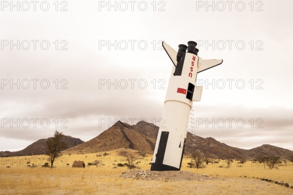 A mock Soviet rocket stands nose-first in the Solitaire, Namibian desert, surrounded by golden grasslands and rugged mountains under cloudy skies