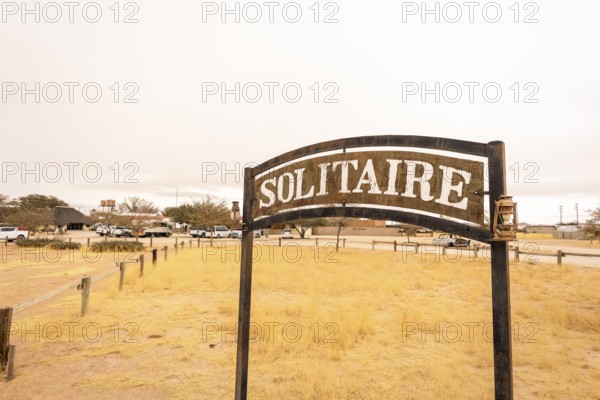 The remote desert settlement of Solitaire in Namibia features a sign displaying the town's name, set against golden sand dunes and rugged mountains
