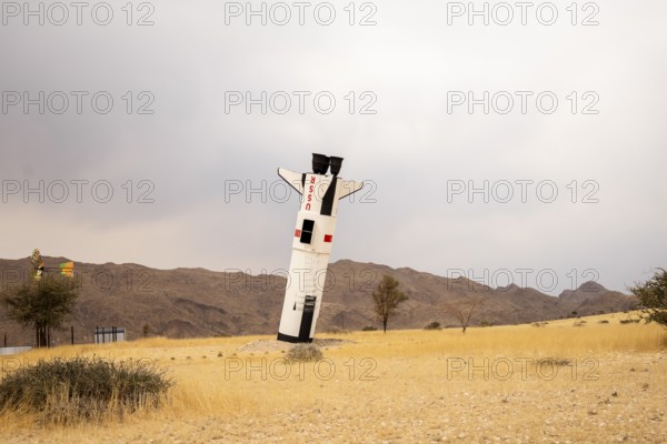 A mock Soviet rocket stands nose-first in the Solitaire, Namibian desert, surrounded by golden grasslands and rugged mountains under cloudy skies