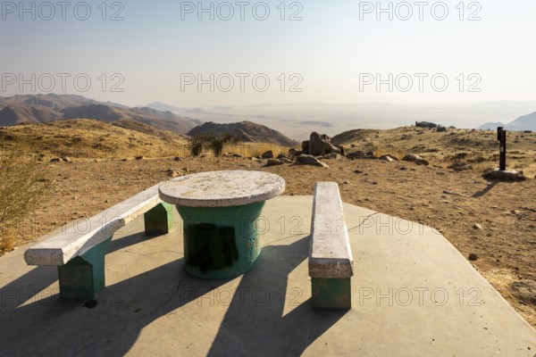 A picnic spot overlooks the vast Namib Desert from Spreetshoogte Pass, with hazy plains, rugged mountains, and a lone house on the ridge under the bright sky