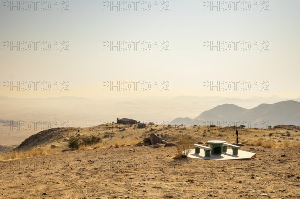A picnic spot overlooks the vast Namib Desert from Spreetshoogte Pass, with hazy plains, rugged mountains, and a lone house on the ridge under the bright sky