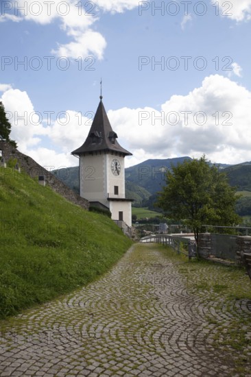 A clock tower at the end of a cobbled path, surrounded by green nature and mountains, Bruck an der Muhr Styria Austria