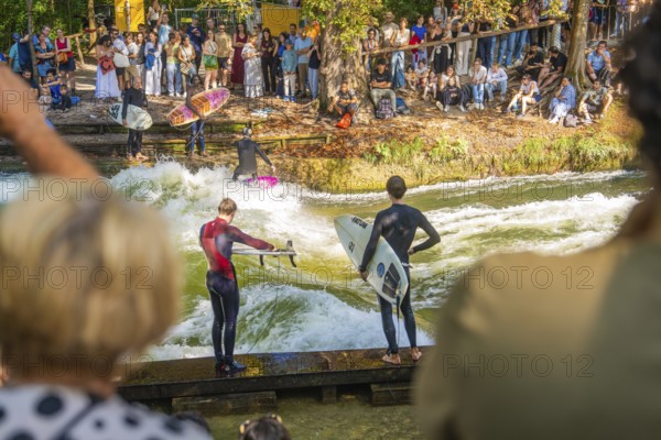 People watching surfers on an artificial wave in a river, surrounded by trees, Munich, Germany