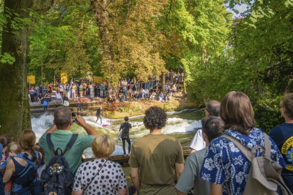 A group of people watching surfers riding in a river, surrounded by trees and summer nature, Munich, Germany