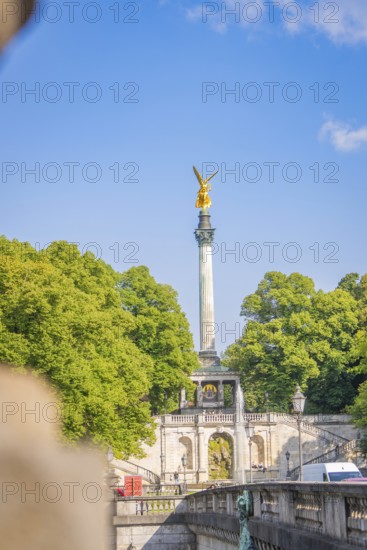 Column statue in golden spire, surrounded by green nature and bright blue sky, Munich, Germany